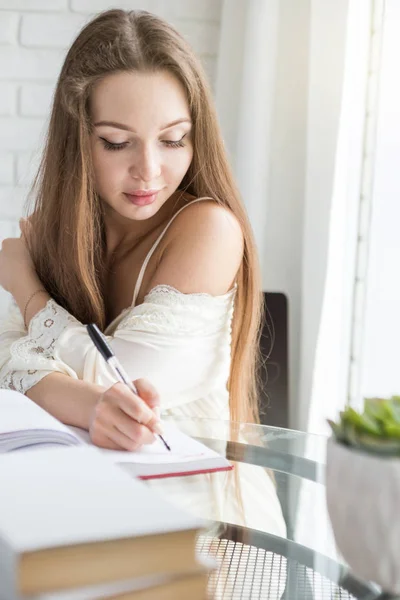 Uma menina bonita em uma camisa de noite está sentada de manhã em uma mesa de vidro e escreve seus pensamentos em um caderno . — Fotografia de Stock