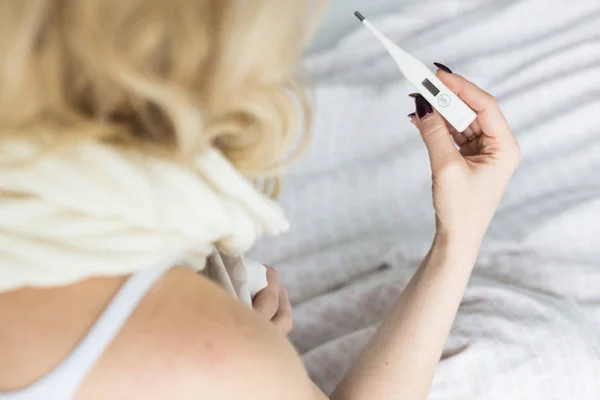 Mooie blonde vrouw zittend in bed met een thermometer en een sjaal gewikkeld rond de nek en een zakdoek. ziekte en gezondheid thuis — Stockfoto