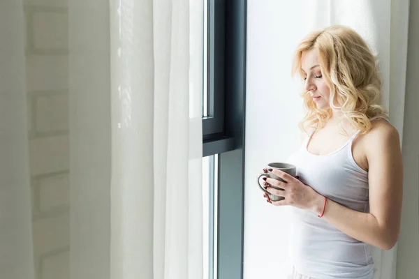 Mulher loira atraente fica perto da janela com uma xícara de café ou chá — Fotografia de Stock