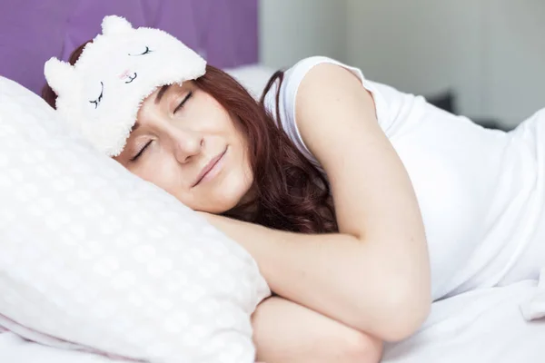 Een aantrekkelijke jonge brunette vrouw wakker in haar bed in een slaap masker. — Stockfoto