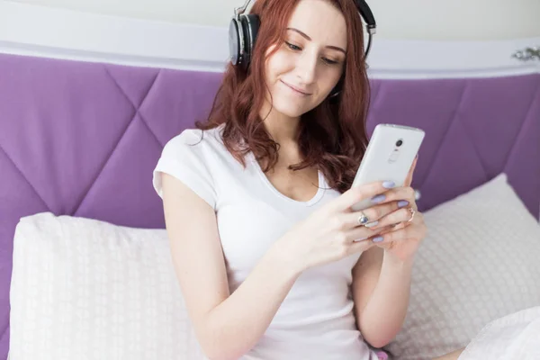 Ελκυστική νεαρή μελαχρινή γυναίκα κάθεται το πρωί στο κρεβάτι της με ακουστικά και ένα smartphone και ακούει μουσική σε καλή διάθεση — Φωτογραφία Αρχείου