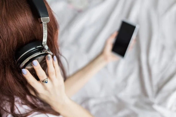 Atractiva joven morena se sienta por la mañana en su cama con auriculares y un teléfono inteligente y escucha música de buen humor — Foto de Stock
