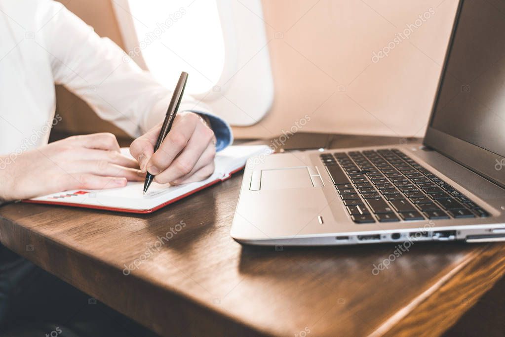 Close-up of a businessmans hand writing in a notebook on the background of a laptop and the interior of an airplane