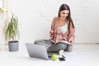 Genç ve mutlu bir kadın parlak bir apartman dairesinde ya da ofis içinde yerde oturuyor ve bir dizüstü bilgisayarda çalışıyor, iş yerinde serbest çalışan bir kız, uzaktan öğrenme öğrencileri, evrak işleri ve çevrimiçi öğrenim kavramı