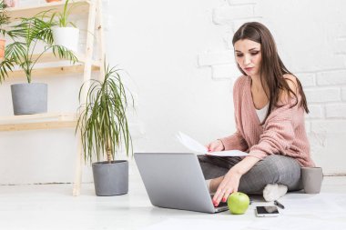 Genç ve mutlu bir kadın parlak bir apartman dairesinde ya da ofis içinde yerde oturuyor ve bir dizüstü bilgisayarda çalışıyor, iş yerinde serbest çalışan bir kız, uzaktan öğrenme öğrencileri, evrak işleri ve çevrimiçi öğrenim kavramı