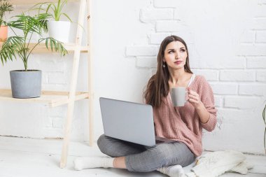 Genç ve mutlu bir kadın parlak bir apartman dairesinde ya da ofis içinde yerde oturuyor ve bir dizüstü bilgisayarda çalışıyor, iş yerinde serbest çalışan bir kız, uzaktan öğrenme öğrencileri, evrak işleri ve çevrimiçi öğrenim kavramı
