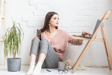 Ev yapımı günlük giysiler içinde mutlu bir kadın parlak bir iç mekanda oturur ve telefon kullanır, kız bir dizüstü bilgisayarda çalışırken ve internette sörf yaparken dinlenir. Rahat serbest çalışma alanı,
