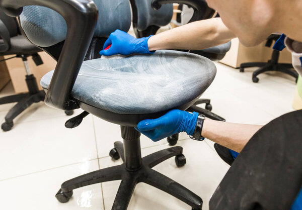 Young man in workwear and rubber gloves cleans the office chair with professional equipment.
