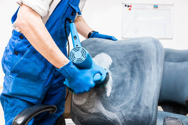 Young man in workwear and rubber gloves cleans the office chair with professional equipment.