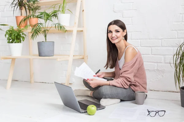 Una joven feliz mujer se sienta en el suelo en un apartamento luminoso o interior de la oficina y trabaja en un ordenador portátil, freelancer chica en el trabajo, el concepto de estudiantes de educación a distancia, papeleo y aprendizaje en línea — Foto de Stock