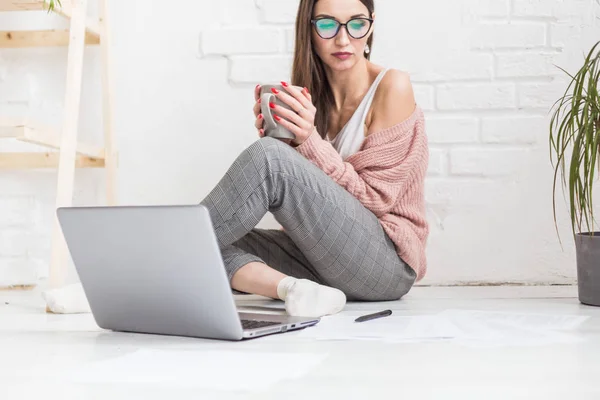 Μια νεαρή ευτυχισμένη γυναίκα κάθεται στο πάτωμα σε ένα φωτεινό διαμέρισμα ή εσωτερικό γραφείο και εργάζεται σε ένα φορητό υπολογιστή, ελεύθερος επαγγελματίας κορίτσι στην εργασία, η έννοια της εξ αποστάσεως μάθησης φοιτητές, γραφειοκρατία και online μάθηση — Φωτογραφία Αρχείου