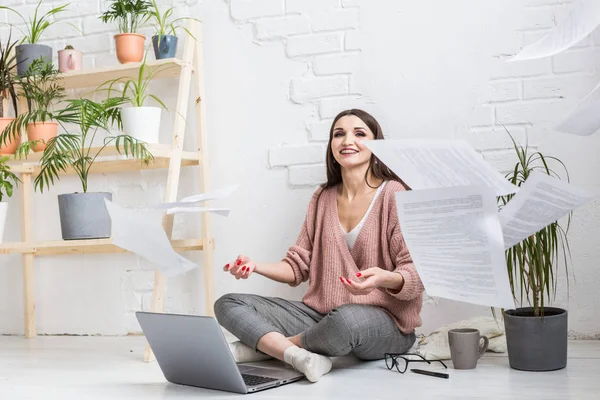Молода щаслива жінка викидає купу паперу і документів, що сидять біля ноутбука, позаштатна дівчина щаслива і в хорошому настрої. хвилювання та натхнення — стокове фото