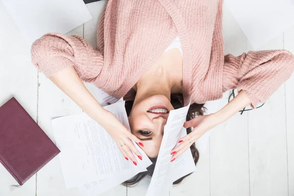Una hermosa mujer yace en el suelo entre papeles y documentos, la chica freelancer sonríe y se relaja durante un descanso del trabajo — Foto de Stock