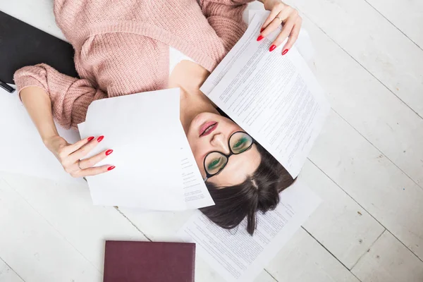 Una hermosa mujer en gafas con estilo se encuentra en el suelo entre los papeles y documentos, la chica freelancer sonríe y se relaja durante un descanso del trabajo — Foto de Stock