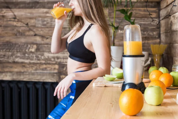Attraktiv tjej i sportkläder förbereder en hälsosam frukost innan träningen i köket, skär frukt och dricker färsk juice gjord i en juiceapparat. Fördelarna med hälsosam kost, fitness och skönhet — Stockfoto