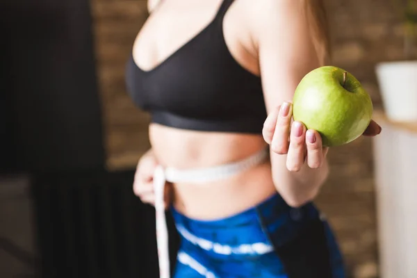 一位穿着运动服装的美丽年轻女子测量测量的飞行腰围, 一手拿着一个苹果。运动、饮食和减肥 — 图库照片