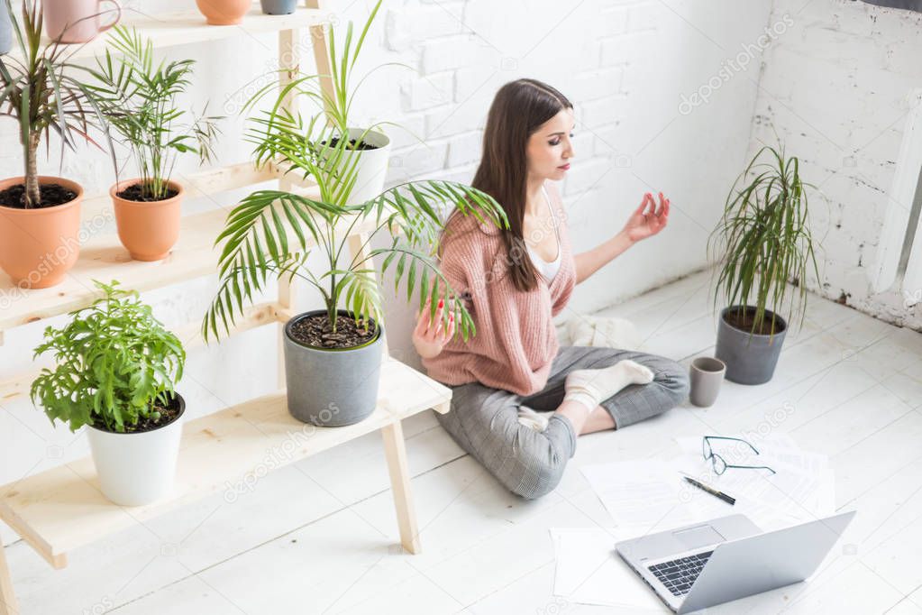 Young happy woman sits on the floor in a yoga pose in a bright apartment and works behind a laptop, a freelancer girl meditates while working. Stress of work