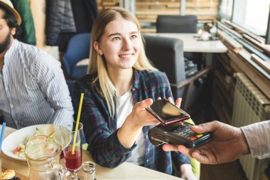 Genç ve çekici bir kadın, kafeteryada bağlantısız akıllı telefon ödemesiyle ödeme yapıyor.