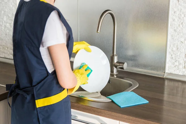 Sarı eldivenli genç bir kadın bulaşıkları lavaboda süngerle yıkıyor. Profesyonel ev temizleme servisi. — Stok fotoğraf