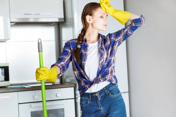 Seorang wanita muda melakukan pembersihan di rumah, mencuci dapur. Ember dengan kain dan sarung tangan kuning di atas meja — Stok Foto