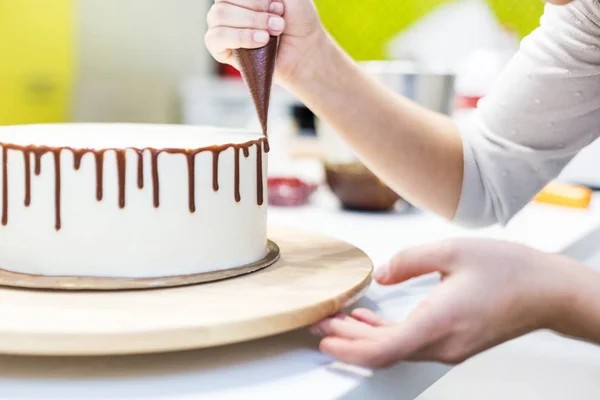 A confectioner squeezes liquid chocolate from a pastry bag onto a white cream biscuit cake on a wooden stand. The concept of homemade pastry, cooking cakes. — Stock Photo, Image