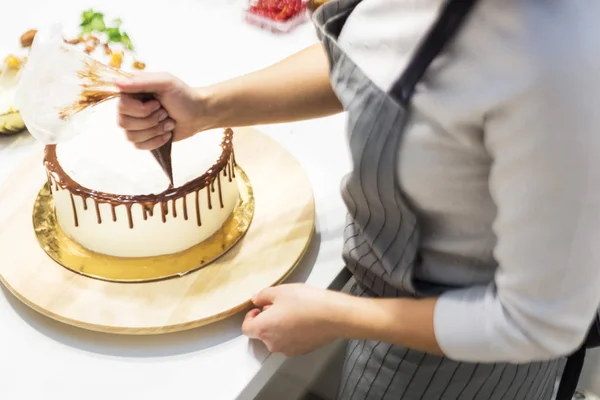 Een banketbakker knijpt vloeibare chocolade uit een gebakje zak op een witte room biscuit cake op een houten tribune. Het concept van zelfgemaakte gebak, koken cakes. — Stockfoto