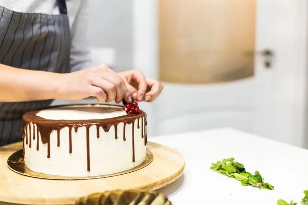 Confectioner dekorerar med bär en kak kaka med vit grädde och choklad. Kakan står på ett trä stativ på ett vitt bord. Konceptet med hembakat bak verk, matlagnings tårtor. — Stockfoto