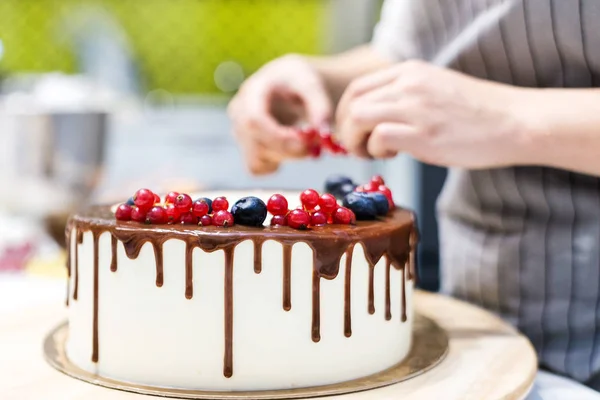 Confectioner dekorerar med bär en kak kaka med vit grädde och choklad. Kakan står på ett trä stativ på ett vitt bord. Konceptet med hembakat bak verk, matlagnings tårtor. — Stockfoto