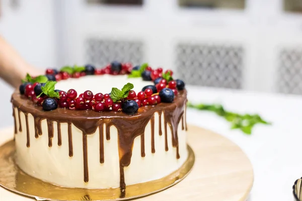 De banketbakkerij versiert met bessen een koekjes cake met witte room en chocolade. Cake staat op een houten stand op een witte tafel. Het concept van zelfgemaakte gebak, koken cakes. — Stockfoto