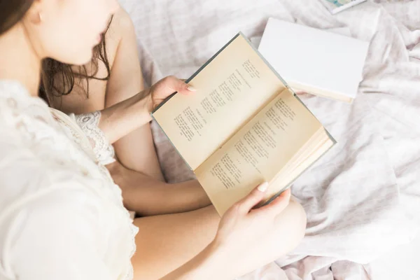 Jovem atraente em uma camisa de noite se senta na cama e lê um livro — Fotografia de Stock