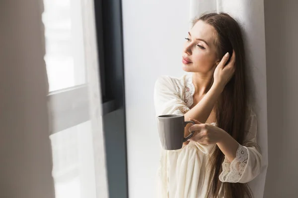 Menina atraente em roupas de noite em pé junto à janela com uma caneca de café — Fotografia de Stock