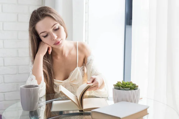 Hermosa joven en un camisón se sienta por la mañana en la mesa de cristal, mira por la ventana y lee un libro — Foto de Stock
