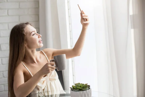 Uma menina bonita em uma camisa de noite senta-se de manhã em uma mesa de vidro, bebe café e tira uma selfie no telefone inicial . — Fotografia de Stock