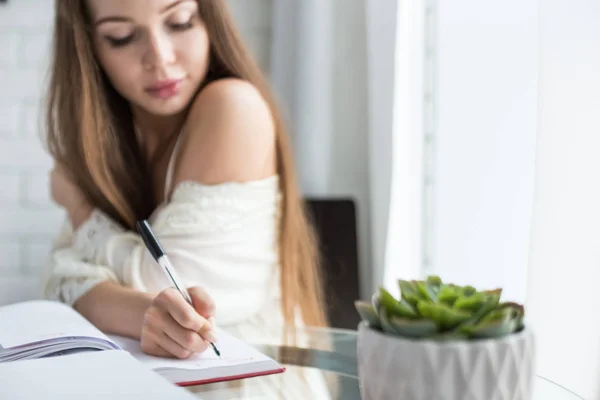 Красива молода дівчина в нічній сорочці сидить вранці за скляним столом і пише свої думки в блокноті . — стокове фото