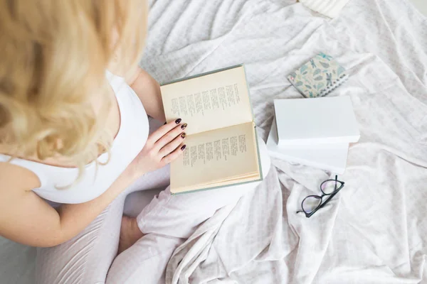 Aantrekkelijke blonde vrouw in bril met zwarte rand zit aan de rand van het bed thuis met een boek en leest — Stockfoto