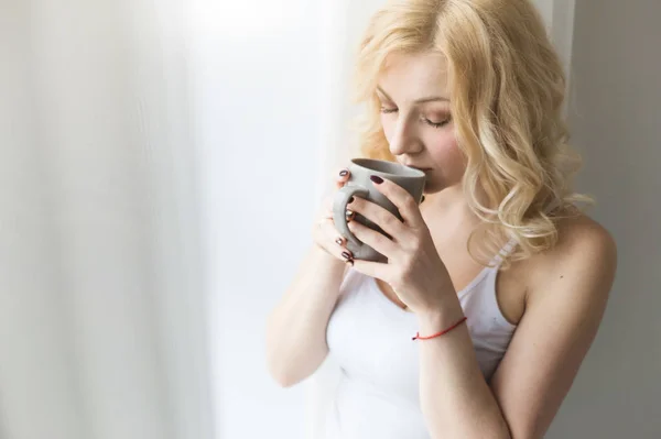 Portret van een aantrekkelijke blonde vrouw die bij het raam staat met een kop koffie of thee — Stockfoto