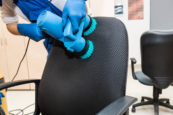 Young man in workwear and rubber gloves cleans the office chair with professional equipment.