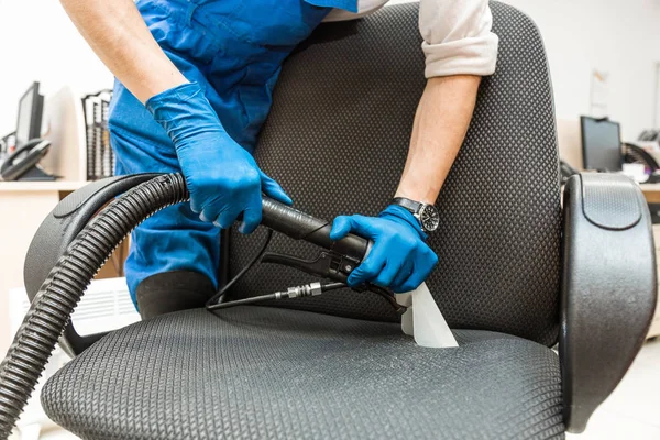 Pemuda dalam pakaian kerja dan sarung tangan karet membersihkan kursi kantor dengan peralatan profesional. — Stok Foto