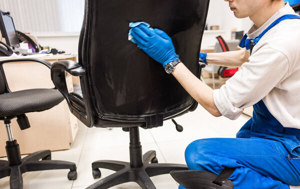 Young man in workwear and rubber gloves cleans the office chair with professional equipment.