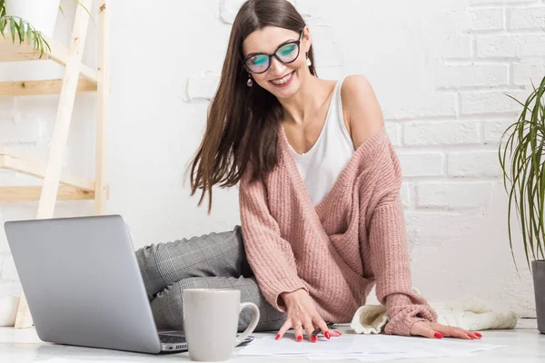 Een jonge vrolijke vrouw zit op de vloer in een licht appartement of kantoorinterieur en werkt op een laptop, freelancer girl op het werk, het concept van afstandsonderwijs studenten, papierwerk en online leren — Stockfoto