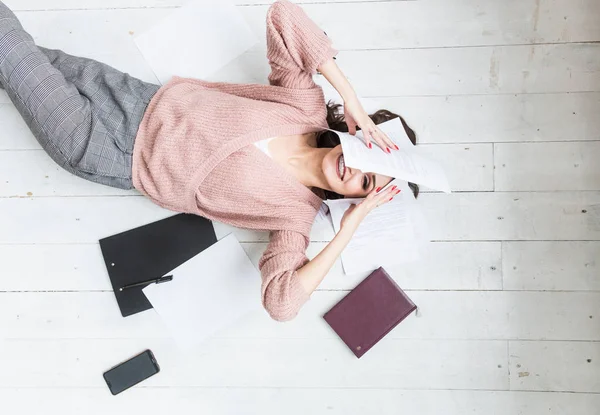 Una hermosa mujer yace en el suelo entre papeles y documentos, la chica freelancer sonríe y se relaja durante un descanso del trabajo — Foto de Stock