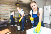 Úklidová služba s profesionálním vybavením během práce. profesionální úklid kuchyňky, chemické čištění pohovky, mytí oken a podlah. muž a ženy v uniformách, kombinézách a gumových rukavicích