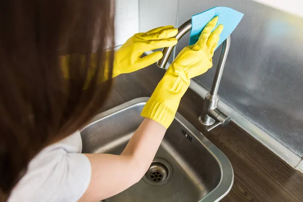 Egy sárga kesztyűs fiatal nő mossa a darut egy szivaccsal a mosogatóban. Háztartási professzionális takarítás — Stock Fotó