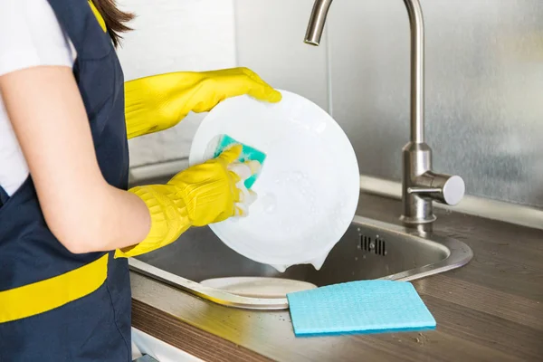 Egy fiatal nő sárga kesztyűben mosogatja a tányérokat szivaccsal a mosogatóban. Háztartási professzionális takarítás. — Stock Fotó