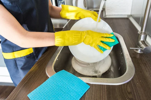 Eine junge Frau in gelben Handschuhen spült Geschirr mit einem Schwamm in der Spüle. Professioneller Reinigungsservice. — Stockfoto