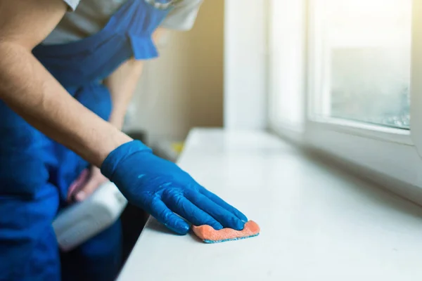 Close-up dari seorang pria berseragam dan sarung tangan biru mencuci jendela dengan scraper jendela. Layanan pembersihan rumah profesional — Stok Foto