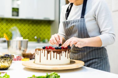 Tatlıcı çilek ile beyaz krem ve çikolata ile bir bisküvi kek dekorlar. Pasta beyaz bir masada ahşap bir stand üzerinde duruyor. Ev yapımı pasta kavramı, yemek pastaları.