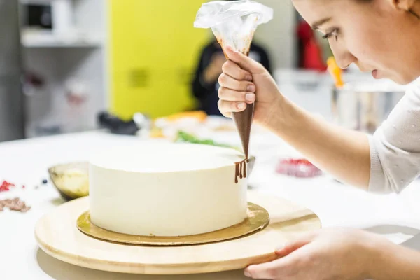 Een banketbakker knijpt vloeibare chocolade uit een gebakje zak op een witte room biscuit cake op een houten tribune. Het concept van zelfgemaakte gebak, koken cakes. — Stockfoto