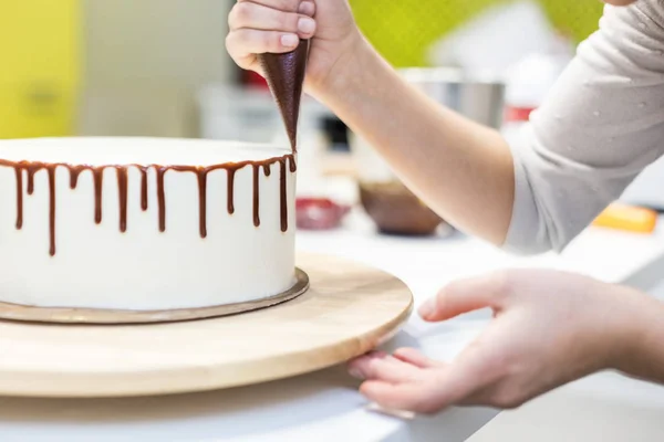 Een banketbakker knijpt vloeibare chocolade uit een gebakje zak op een witte room biscuit cake op een houten tribune. Het concept van zelfgemaakte gebak, koken cakes. — Stockfoto