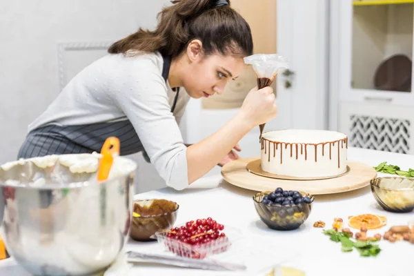 Een banketbakker knijpt vloeibare chocolade uit een gebakje zak op een witte room biscuit cake op een houten tribune. Het concept van zelfgemaakte gebak, koken cakes. — Stockfoto